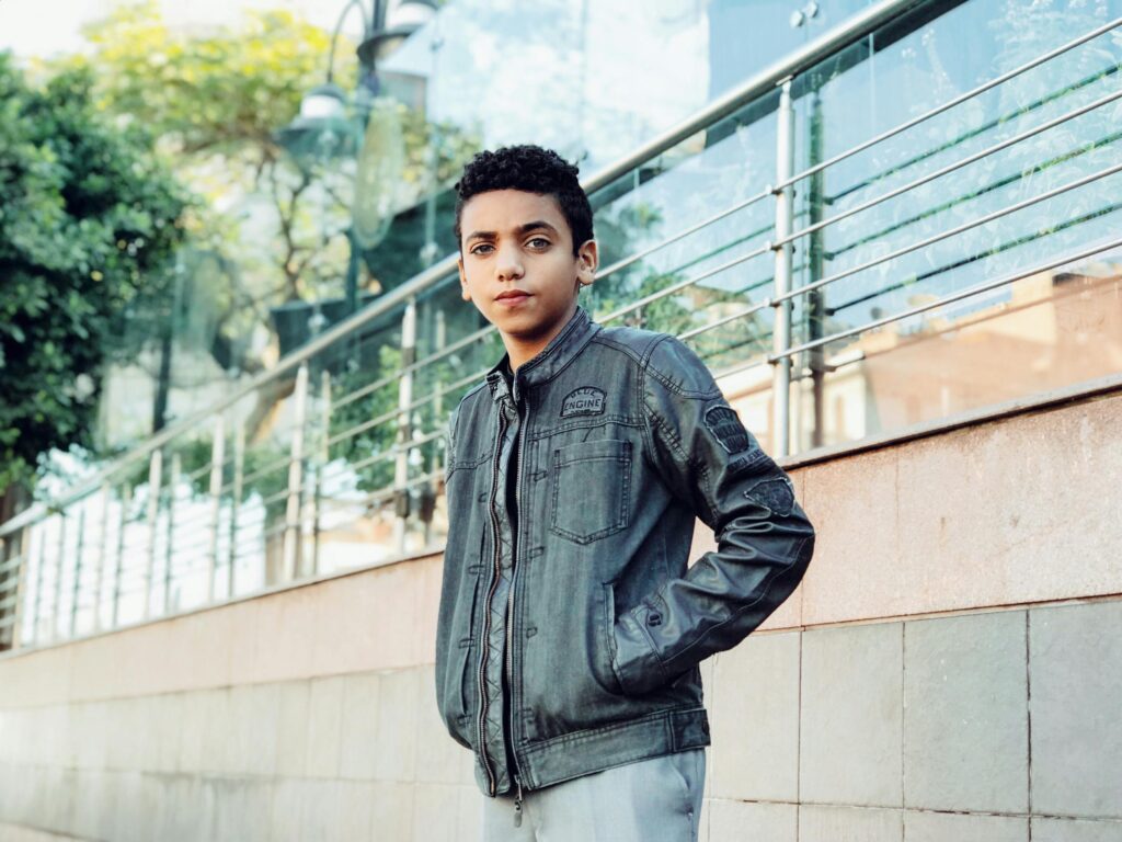 Stylish teenage boy in a jacket poses confidently outdoors against city background.